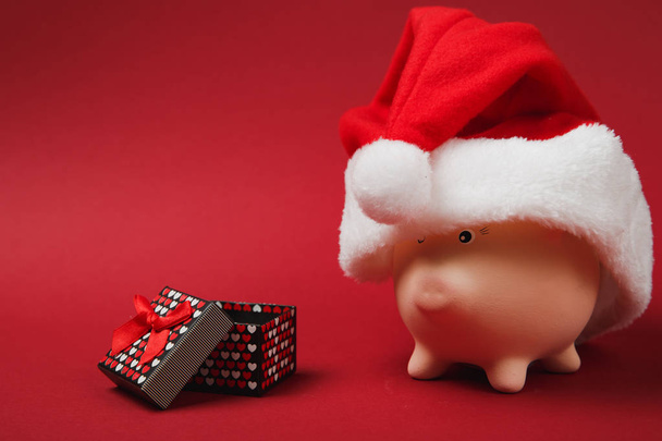 Pink piggy money bank with Christmas hat, present box with gift ribbon isolated on red background. Money accumulation, investment, wealth concept. Happy New Year, birthday holiday. Mock up copy space - Valokuva, kuva