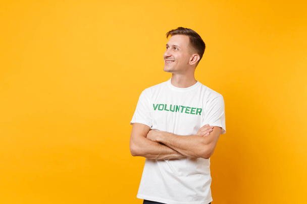 Portrait of happy smiling satisfied young man in white t-shirt with written inscription green title volunteer isolated on yellow background. Voluntary free assistance help, charity grace work concept - Zdjęcie, obraz