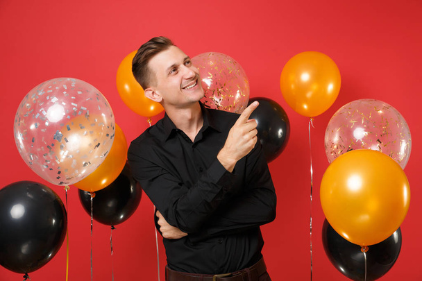 Joyful young man in black classic shirt looking up pointing index finger aside on bright red background air balloons. St. Valentine's, Women's Day Happy New Year birthday mockup holiday party concept - Photo, Image