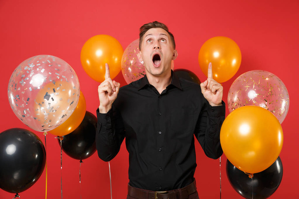 Shocked young man in classic shirt looking up, pointing index fingers up on red background air balloons. St. Valentine's International Women's Day Happy New Year birthday mockup holiday party concept - Photo, Image
