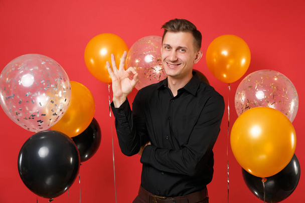 Lächelnder junger Mann in schwarzem klassischen Hemd feiert und zeigt ein Ok-Zeichen auf rotem Hintergrund Luftballons. St. Valentin, internationaler Frauentag, frohes neues Jahr, Geburtstagsparty-Konzept - Foto, Bild