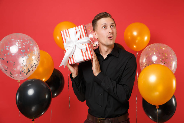 Ohromen, mladý muž v černé klasické tričko drží červený rámeček s darem, na jasně červeném pozadí s vrtulovým pohonem. Svatý Valentýn, žen den, šťastný nový rok, narozeniny maketa holiday party koncept - Fotografie, Obrázek
