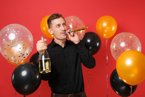 Grappige jongeman in zwarte klassieke shirt met het afspelen van de pijp houdt fles champagne op rode achtergrond lucht ballonnen. Concept vakantie feest van St. Valentijnsdag, de dag van de vrouw, Happy New Year verjaardag mockup - Foto, afbeelding