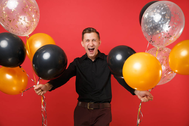 Happy young man in black classic shirt holding air balloons, celebrating isolated on bright red background. Valentine's, International Women's Day Happy New Year birthday mockup holiday party concept - Zdjęcie, obraz