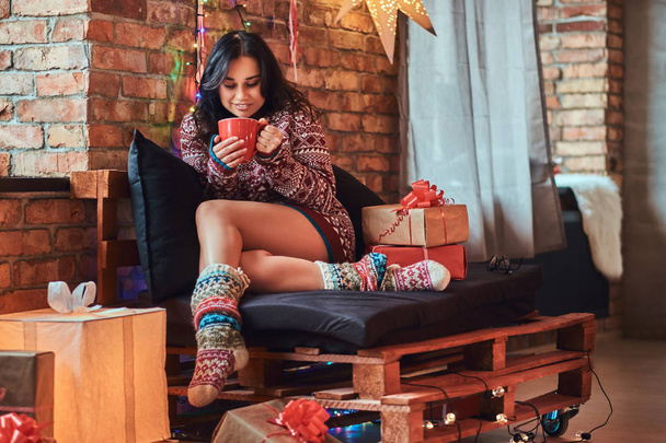 Hermosa chica calentándose con una taza de café, disfrutando de la mañana de Navidad mientras se sienta en un sofá en una habitación decorada con interior loft
. - Foto, imagen