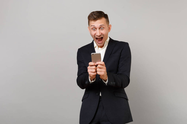 Surprised happy young business man screaming keeping mouth open wide, using mobile phone typing sms message isolated on grey background. Achievement career wealth business concept. Mock up copy space - Foto, Imagem