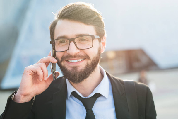 Portrait of smiling bearded businessman in suit using smartphone on office build and sunlight background. - Foto, imagen