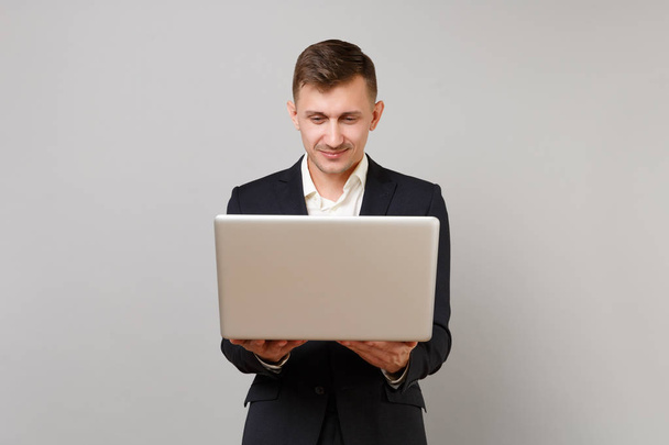 Aantrekkelijke jonge zakenman in klassiek zwart pak en hemd werkt op laptop PC geïsoleerd op grijs muur achtergrond in de studio. Prestatie carrière rijkdom bedrijfsconcept. Bespotten kopie databaseruimte - Foto, afbeelding