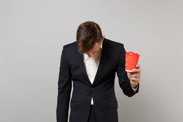 Exhausted young business man in classic black suit with lowered head holding paper cup with coffee or tea isolated on grey background. Achievement career wealth business concept. Mock up copy space - Foto, Imagem