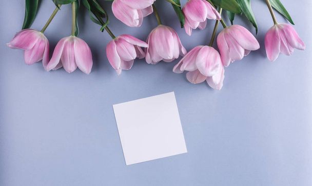 rosa Tulpenblüten und ein Blatt Papier auf hellblauem Hintergrund. Grußkarte oder Hochzeitseinladung. flache Lage, Draufsicht, Kopierraum - Foto, Bild