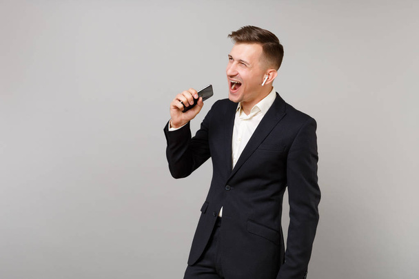 Веселый молодой бизнесмен в черном костюме с беспроводными наушниками, слушая музыку, держит мобильный телефон изолированным на сером фоне. Достижение карьерного богатства бизнес-концепции. Пространство для копирования
 - Фото, изображение