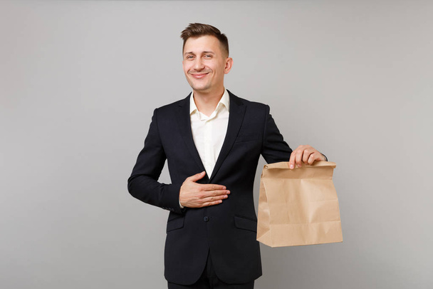 Joyful business man holding brown clear empty blank craft paper bag for takeaway mock up isolated on grey background. Achievement career wealth business concept. Food products delivery to work office - Fotó, kép