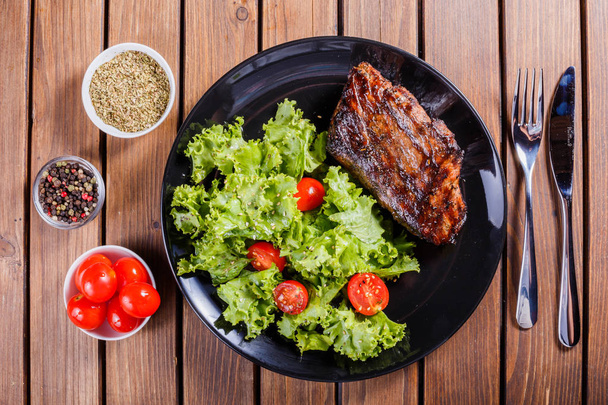 Steak de porc grillé avec salade de légumes frais et tomates sur assiette noire, fond en bois
 - Photo, image