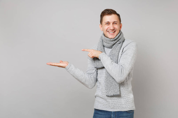 fröhlicher junger Mann im grauen Pullover, Schal mit erhobenem Zeigefinger, die Hand zur Seite auf grauem Hintergrund. Gesunder Lebensstil, aufrichtige Emotionen, Konzept für die kalte Jahreszeit. Attrappe Kopierraum - Foto, Bild