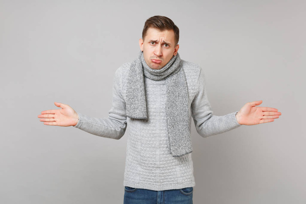 Offended young man in gray sweater, scarf pouting lips, spreading hands isolated on grey wall background. Healthy fashion lifestyle, people sincere emotions, cold season concept. Mock up copy space - Foto, imagen