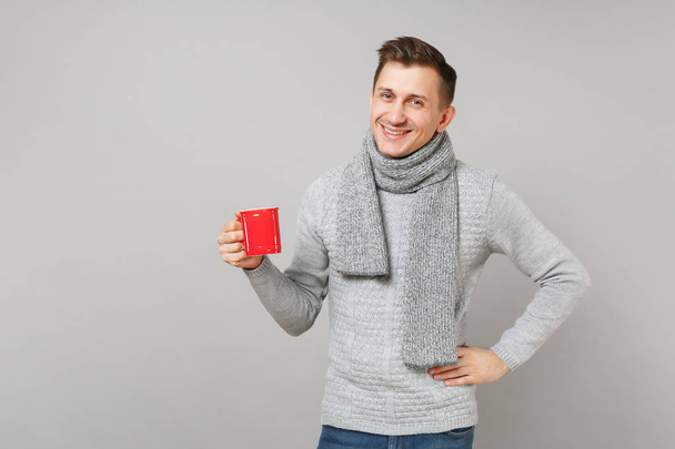 fröhlicher junger Mann in grauem Pullover, Schal mit roter Tasse Kaffee oder Tee auf grauem Hintergrund im Studio. Gesunder Lebensstil, Krankheitsbehandlung, Konzept der kalten Jahreszeit. Attrappe Kopierraum - Foto, Bild