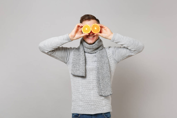 Lächelnder junger Mann in grauem Pullover, Schal, der die Augen mit Orangen auf grauem Hintergrund im Atelier verdeckt. Gesunder Lebensstil, aufrichtige Emotionen, Konzept für die kalte Jahreszeit. Attrappe Kopierraum - Foto, Bild