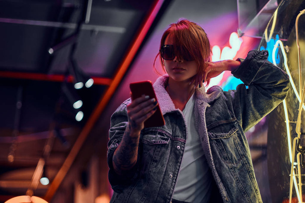 Élégante rousse fille à l'aide d'un téléphone tout en s'appuyant sur le panneau lumineux dans la nuit dans la rue
. - Photo, image
