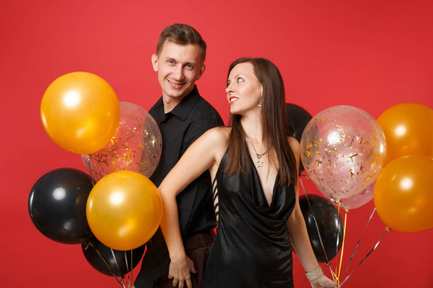 Εντυπωσιακό νεαρό ζευγάρι σε μαύρα ρούχα γιορτάζουν γενέθλια γιορτή απομονώνονται σε λαμπερό κόκκινο φόντο και αερόστατα. Έννοια της γυναίκας του Αγίου Βαλεντίνου ημέρα ευτυχισμένο το νέο έτος 2019. - Φωτογραφία, εικόνα