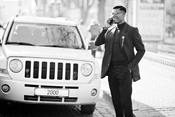 Amazingly looking african american man wear at blue blazer with brooch, black turtleneck and glasses posed at street. Fashionable black guy speaking on phone against car. - Foto, imagen