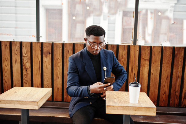 Amazingly looking african american man wear at blue blazer with brooch, black turtleneck and glasses posed at street. Fashionable black guy with cup of coffe looking at his phone. - Zdjęcie, obraz