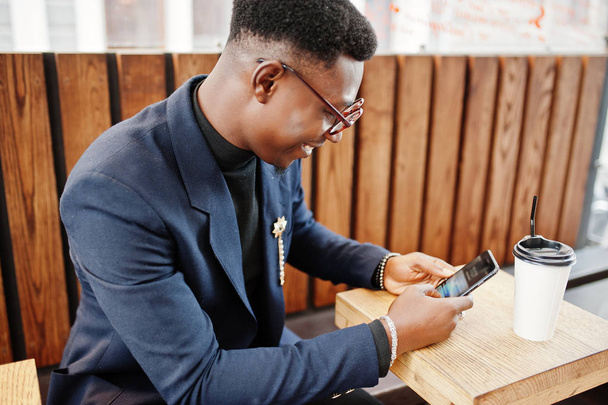 Amazingly looking african american man wear at blue blazer with brooch, black turtleneck and glasses posed at street. Fashionable black guy with cup of coffe looking at his phone. - Valokuva, kuva