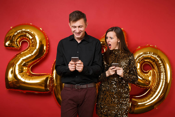 Couple guy girl in dress black shirt celebrating holiday party hold cellphone isolated on bright red wall background golden numbers air balloons studio portrait. Happy New Year 2019 Christmas concept - Photo, Image