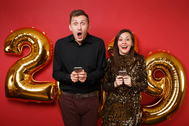 Couple guy girl in dress black shirt celebrating holiday party hold cellphone isolated on bright red wall background golden numbers air balloons studio portrait. Happy New Year 2019 Christmas concept - Foto, Bild