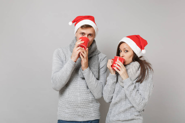Fun couple girl guy in red Santa Christmas hat gray sweaters scarves hold cups of tea isolated on grey wall background, studio portrait. Happy New Year 2019 holiday party concept. Mock up copy space - Foto, immagini