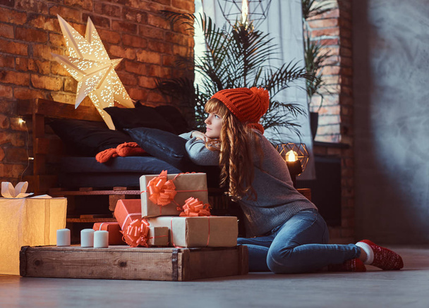 Рыжая девушка, сидящая на полу, мечтательно смотрит в окно в украшенную гостиную на Рождество
. - Фото, изображение