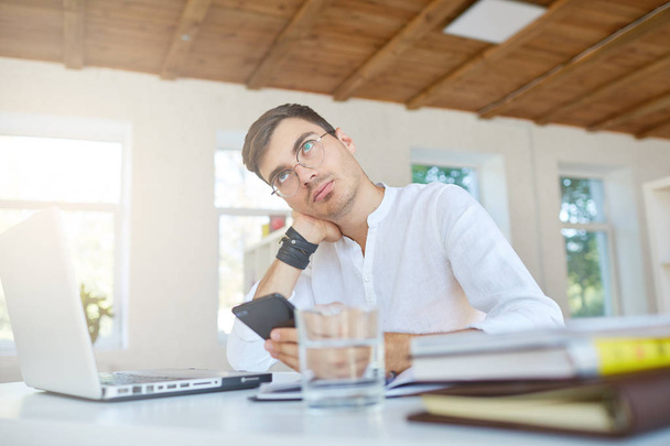 Портрет вдумчивого привлекательного молодого бизнесмена носит белую рубашку и очки, используя ноутбук и смартфон, работающие за столом в офисе
 - Фото, изображение