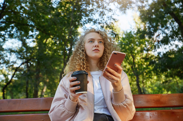 Genç güzel kız sarışın kahve içme bir bankta oturur, biriyle konuşan bir arkadaşı üzerinde görünüyor, telefonu elinde tutan, aşağıdan, park arka plan okuma messageview düşünen. - Fotoğraf, Görsel