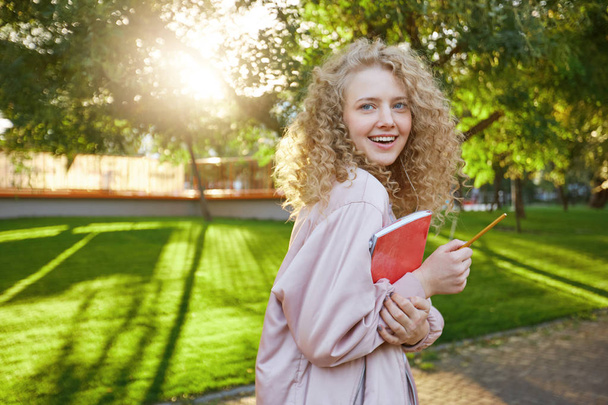 Νεαρή ξανθιά μαλλιά φοιτήτρια βόλτες μέσα από το πάρκο, με ένα κόκκινο σημειωματάριο και ένα μολύβι στα χέρια, ακουστικά στα αυτιά της, με την αγαπημένη μουσική, εξετάζει τη φωτογραφική μηχανή και χαμόγελα - Φωτογραφία, εικόνα