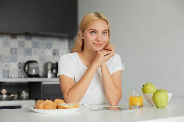 Photo of young blonde girl sits at the kitchen table in the morning, smiling, looks glad to the window, on the table -a glass of juice, apples, buns, dressed in a domestic T-shirt, kitchen background - Фото, зображення