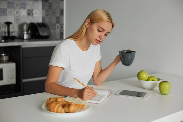 Young blonde girl planning her day, keeps a big grey cup, writes plans in a dairy. Croissants on a plate, apples, are there on a white kitchen table, kitchen background - Photo, Image