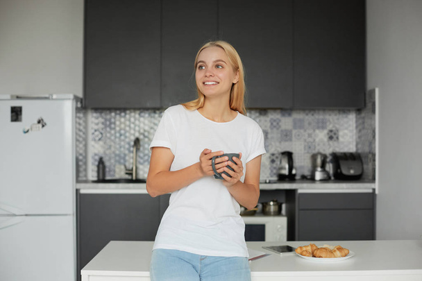Happy young blonde girl in a good mood stands leans on the table, smiling, having breakfast, keeps a big grey teacup, looking away, dreaming, thinking about the coming day, on a kitchen background - Photo, Image