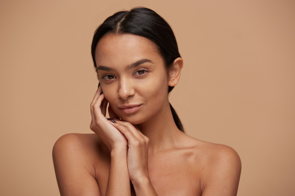 Head shot of an attractive mixed race girl, looking gently and tenderly, demonstrates healthy beautiful skin, calm and pleased, keeps hands near the face, over beige background - Фото, зображення