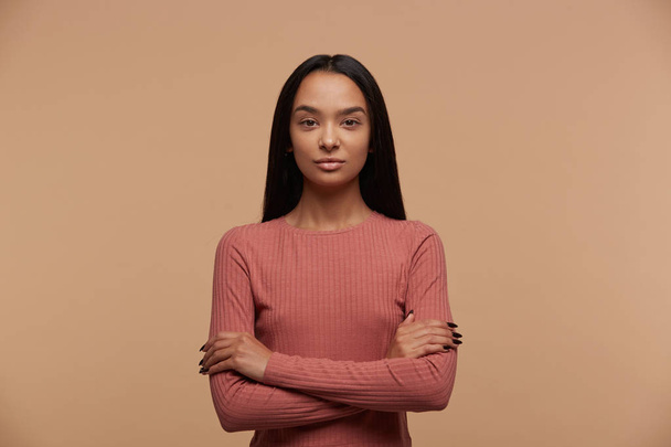 Portrait of calm, confident, attractive young mixed race brunette girl stands with arms crossed, dressed casual, on beige background - Fotó, kép
