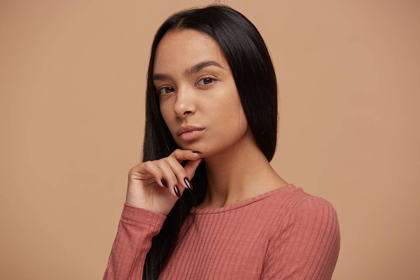 Attractive young woman looks puzzled, trying to remember something important, looks camera on keep one hand on a chin, thoughtful, wears casual clouth, on a on beige background - Foto, afbeelding