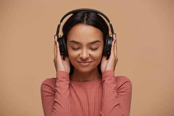 Portrait of a young mixedrace girl listens to her favorite music in big black headphones, closing her eyes from pleasure, enjoyment, music lover, likes lyric songs about love, on beige background - Photo, Image