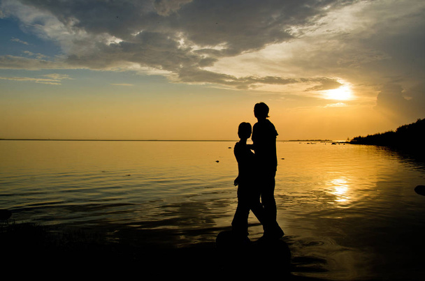 ζευγάρι των εραστών στην ακτή θαυμάζει το ηλιοβασίλεμα πάνω από το νερό - Φωτογραφία, εικόνα