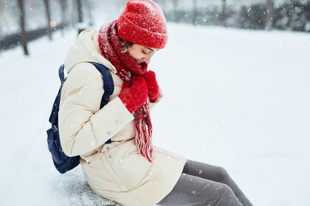 Wesoły uśmiechnięta kobieta w białym dół kurtki i czerwoną czapkę, szalik i rękawiczki siedzi na ławce w alei na Snowy ulicy po Zamieć w mieście. zimowe miasto po śnieżyce i śniegu. - Zdjęcie, obraz