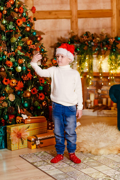 a seven-year-old boy in Santa's cap with a phone in his hands stands near a large elegant Christmas tree. A child takes a selfie - Φωτογραφία, εικόνα