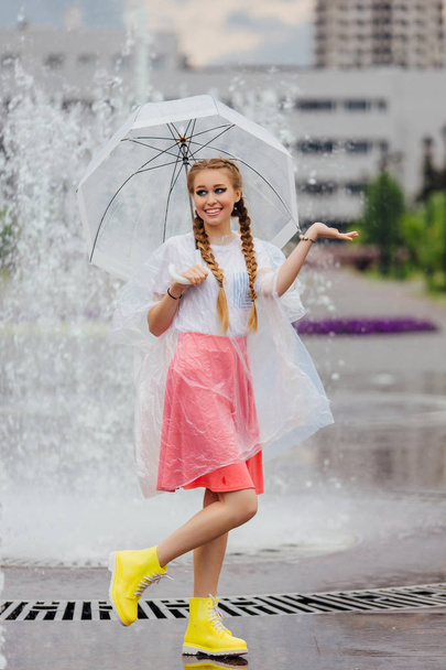 Όμορφη κοπέλα με δύο πλεξούδες σε κίτρινες μπότες και με διάφανη ομπρέλα στέκεται κοντά σε σιντριβάνι. Βροχερή μέρα στη πόλη. - Φωτογραφία, εικόνα