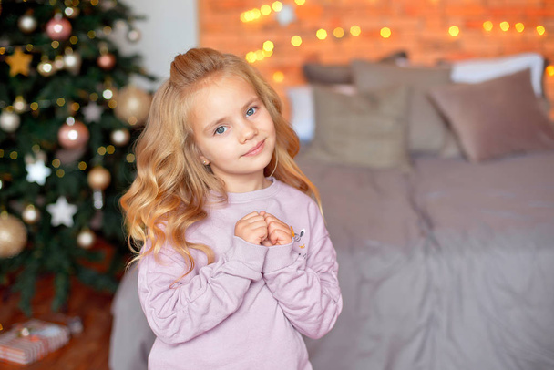 Beautiful girl with blond curls in anticipation of gifts in the background Christmas lights and bed . New Year and Christmas concept. - Fotoğraf, Görsel