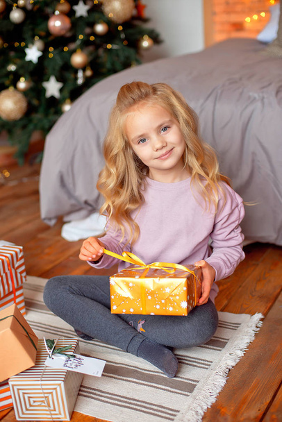 クリスマスの朝、ブロンドの髪とパジャマの女の子がベッドの近くに座っていると、クリスマス ツリーの背景に贈り物を見てします。新年、クリスマス コンセプト. - 写真・画像