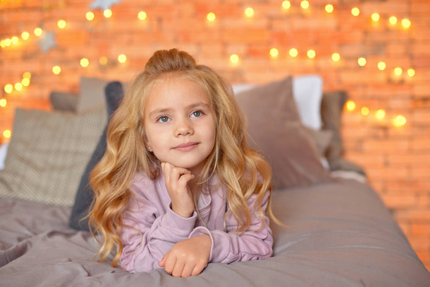 Beautiful girl with blond curls in anticipation of gifts in the background Christmas lights and bed . New Year and Christmas concept. - Photo, image