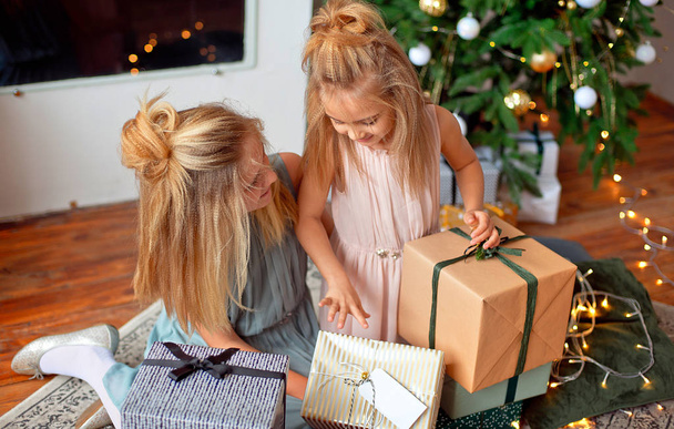 Two sisters with blond hair in luxurious dresses are sitting near the Christmas tree and looking at the gifts. New Year and Christmas Concept. - Foto, imagen