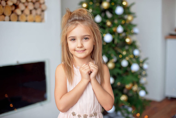 Happy little girl in luxury dress holds hands near face.With colorful lights and Christmas tree on background. Holidays, christmas, new year, x-mas concep - Photo, image