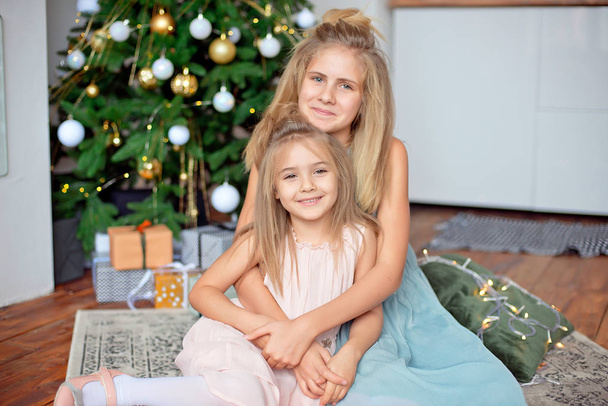 金髪の 2 人の姉妹は、クリスマス ツリーの背景に座っています。新年とクリスマスのコンセプト. - 写真・画像